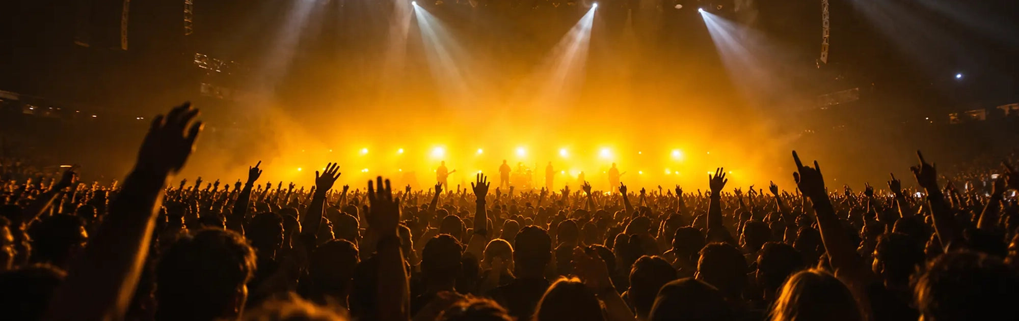A concert crowd with raised hands in a large venue, illuminated by bright yellow and orange stage lights with white spotlights shining down.