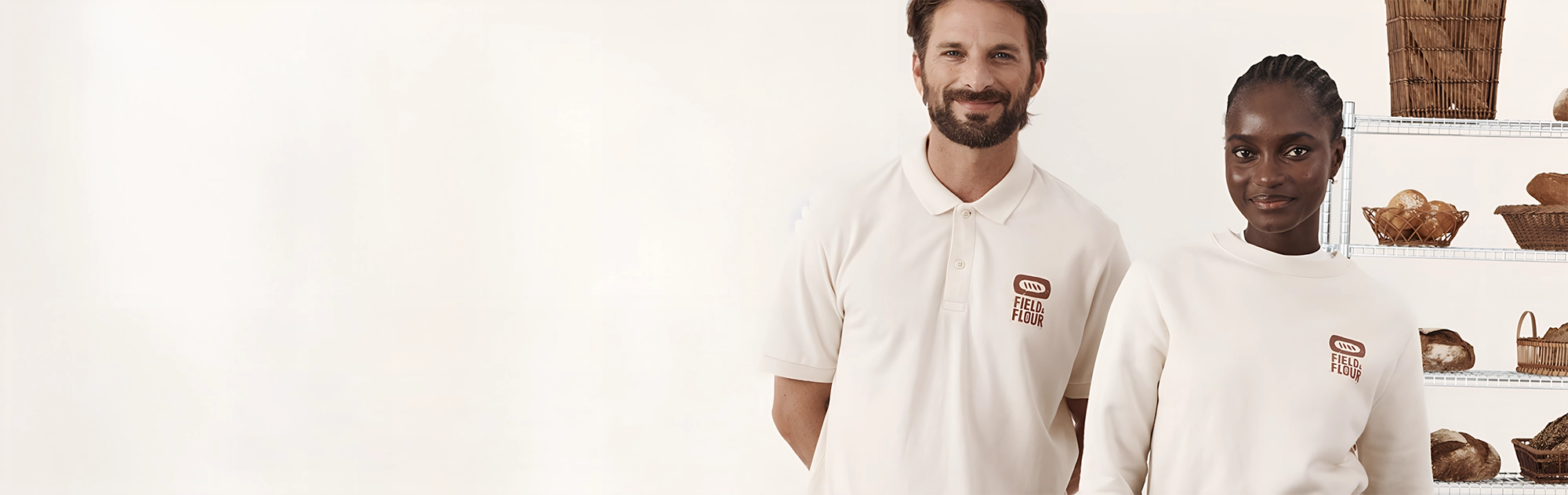 Two people wearing matching white polo shirts with a small printed red logo on the left chest, standing in front of a white wall with shelves of bread baskets on the right.