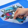 Hand peeling a colourful shark graphic DTF from a sheet on a blue surface