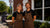 A smiling man and woman standing side by side in front of a restaurant storefront, both wearing matching brown aprons with the brand name 'BOLD STREET HOUSE' printed in white. The woman wears a black shirt and the man wears a black polo, set against a sunny outdoor patio and brick-and-glass building background.