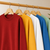 A row of crewneck sweatshirts in red, mustard yellow, white, blue, and sage green, hanging on wooden hangers from a light wood clothing rack.
