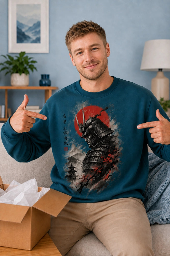 A smiling man wearing a teal long-sleeve sweatshirt with a large samurai graphic points to the chest design while sitting on a couch in a living room. An open cardboard box with tissue paper is visible in the foreground.