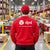 A man viewed from behind wearing a red DPD delivery uniform sweatshirt and matching red cap in a warehouse setting with conveyor belts and cardboard boxes.