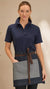 A woman wearing a navy blue polo shirt and a matching apron with a gray front pocket and brown belt tie, standing against a plain beige background.