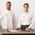 Two hotel staff members wearing matching white long-sleeve collared shirts with 'NIMLES' embroidered on the chest, standing behind a reception desk with a service bell and a booklet.