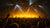 A concert stage filled with bright yellow and white spotlights, with a large crowd of people raising their hands in the foreground.