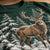 A close-up of a dark green sweatshirt featuring a detailed graphic print of a brown deer with large antlers walking through a snowy pine forest.