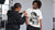 A man wears a white graphic T-shirt with a black-and-grey photo print and a small orange accent, while a woman in a black hoodie adjusts her collar.