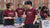 Four students wearing matching maroon t-shirts with a white 'Music Society Liverpool' logo stand behind a booth in front of a red brick university building. One student holds a brochure, and a sign on the right reads 'Music Society Liverpool'.