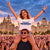 A woman wearing a white graphic T-shirt that reads 'ON THE WATERFRONT' and 'FIRST CLASS, LIVERPOOL' is joyfully sitting on a man's shoulders in a crowded outdoor concert setting at dusk, with Liverpool landmarks in the background.