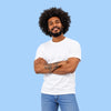 A man with an afro hairstyle, beard, and tattoos wearing a plain white t-shirt and blue jeans, standing with arms crossed against a solid light blue background.